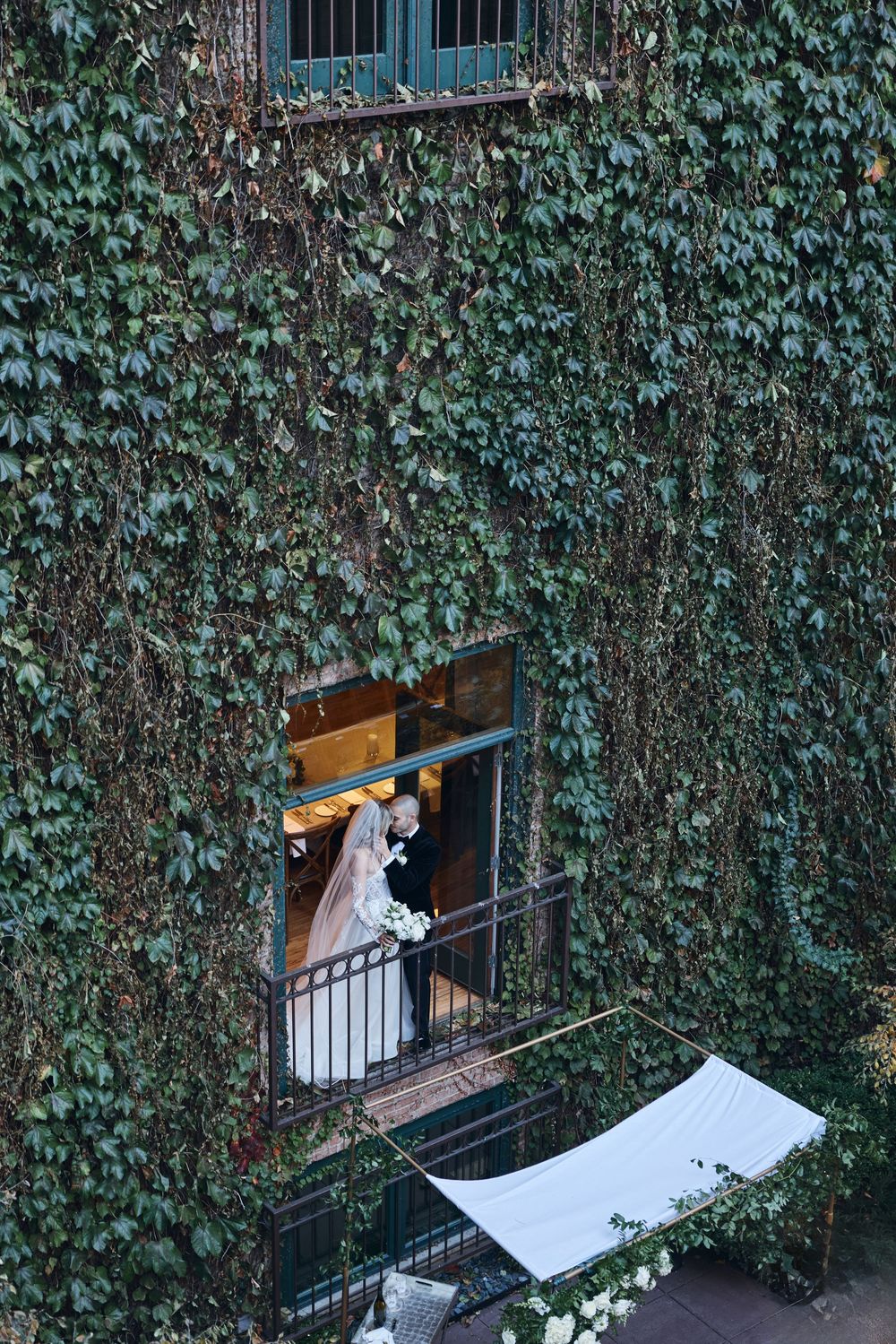 A glimpse through an ivy-covered wall shows a wedding dress through a balcony doorway.