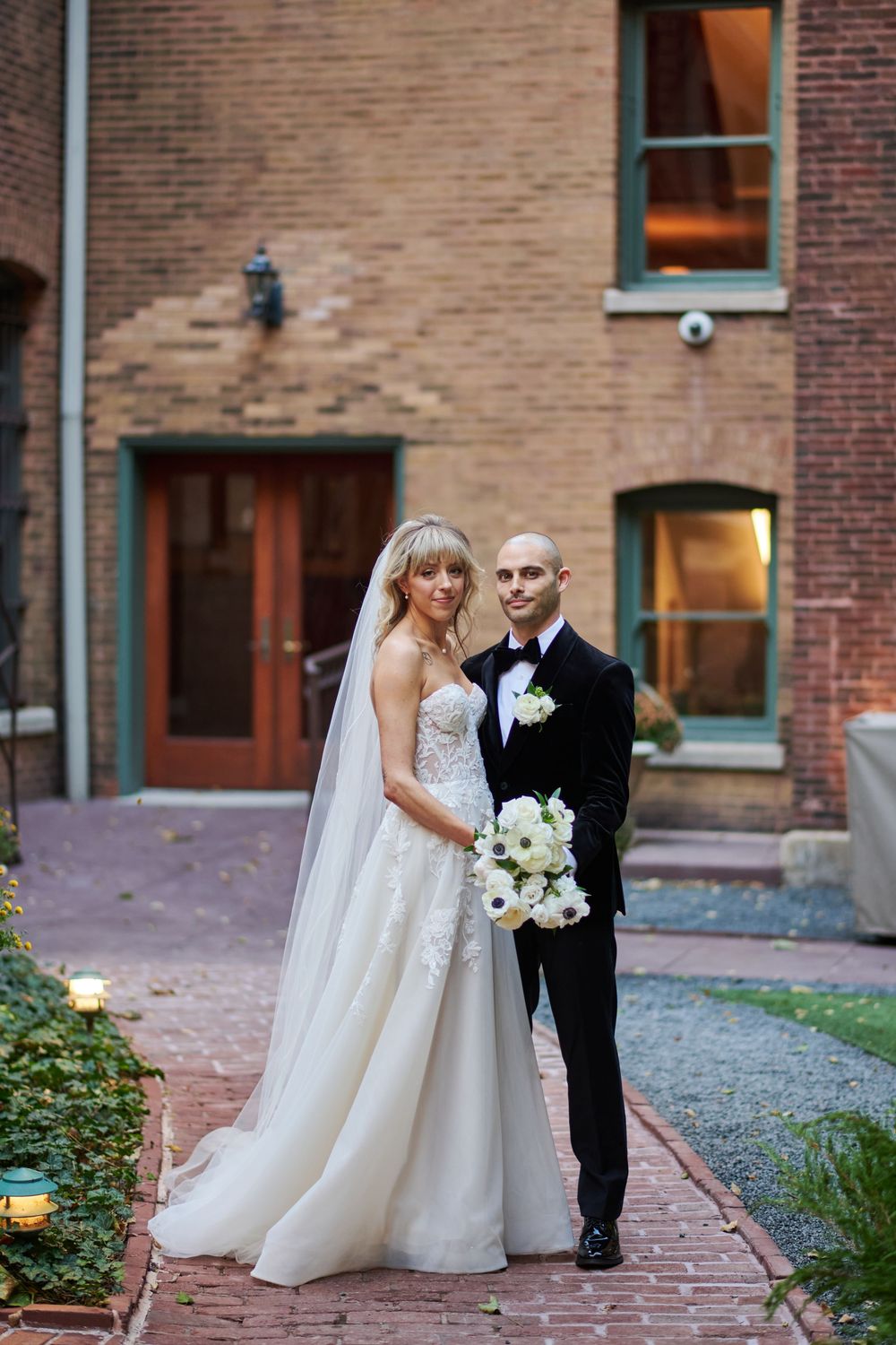 Wedding couple poses for romantic portraits against a brick building backdrop with beautiful landscaping.