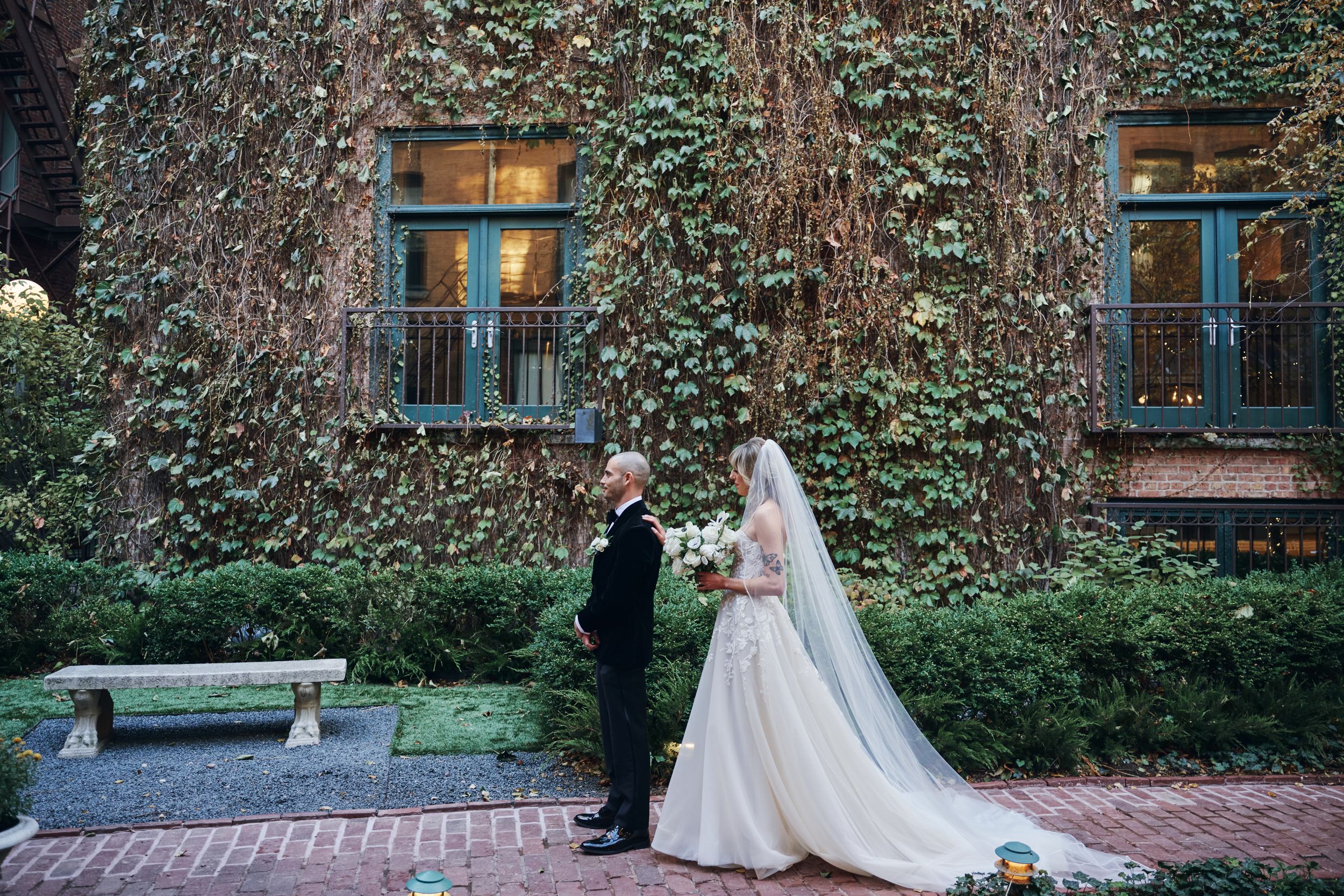Elegant wedding couple poses against ivy-covered brick wall with glowing windows in romantic garden setting.