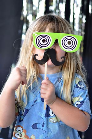 Child wearing a blue shirt holding up silly cartoon glasses and mustache props.