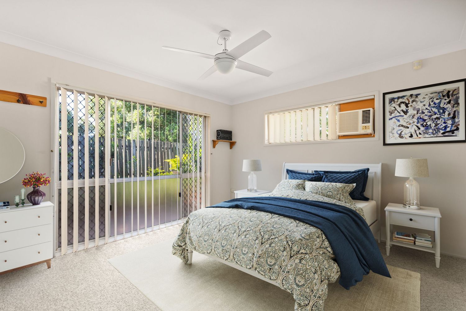 Bright master bedroom with blue bedding, ceiling fan, and sliding glass doors opening to a lush garden.