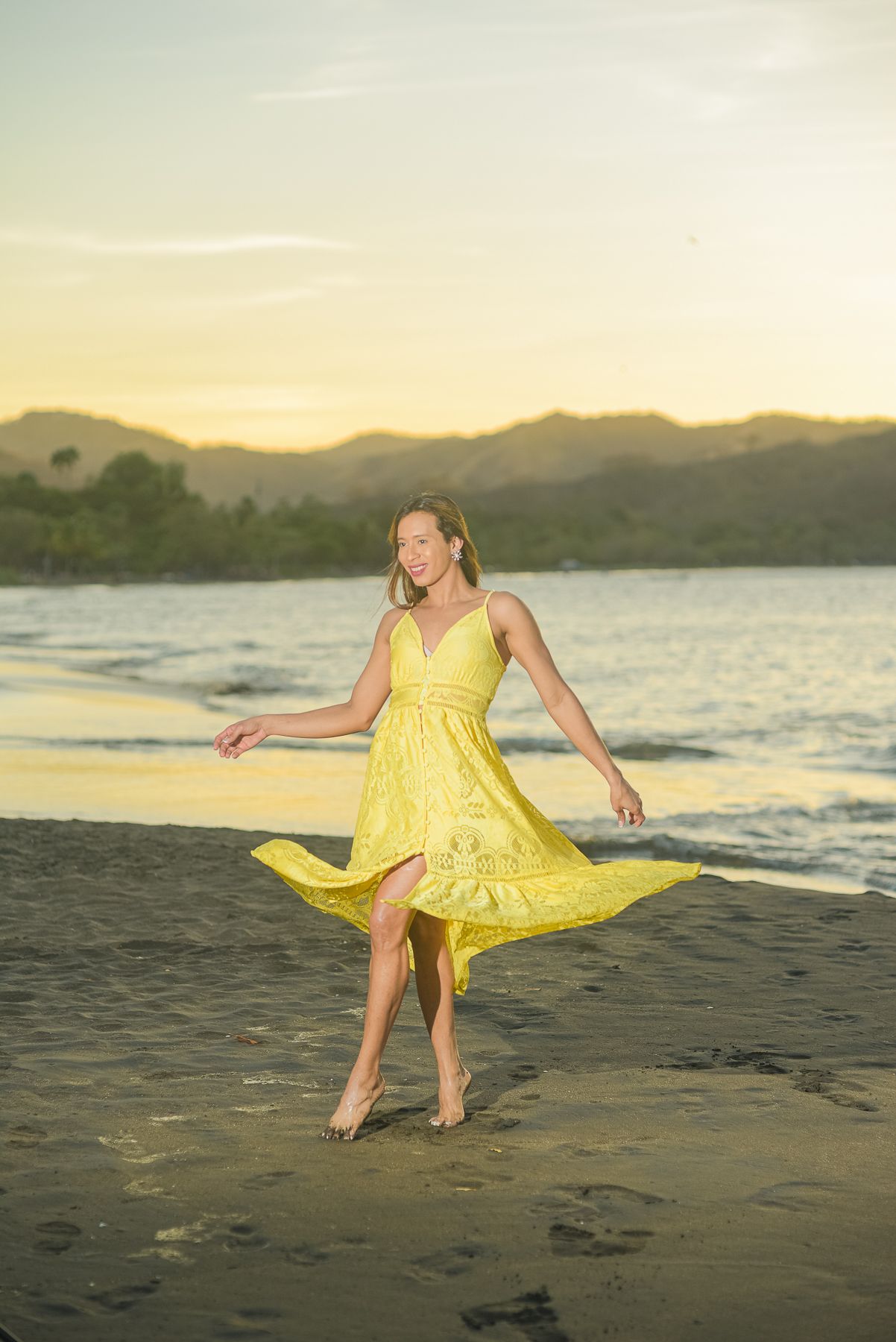 A person in a yellow sundress walks along a scenic beach at sunset with mountains in the background.