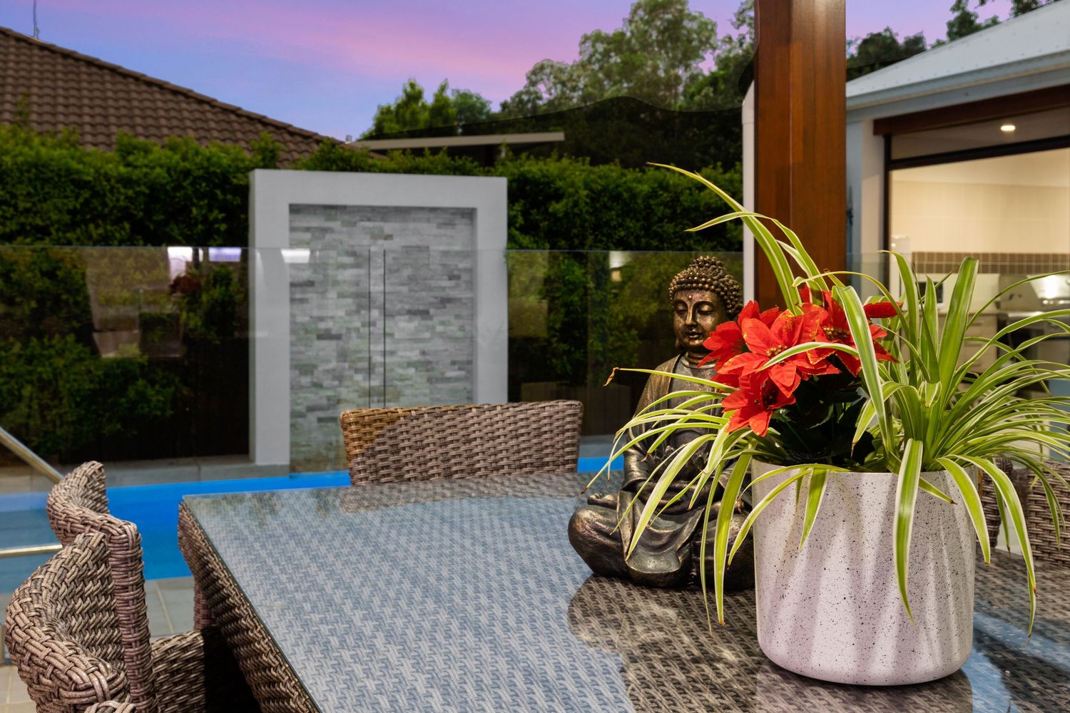 Outdoor patio with wicker furniture, potted plants, Buddha statue, and water feature near pool at dusk.