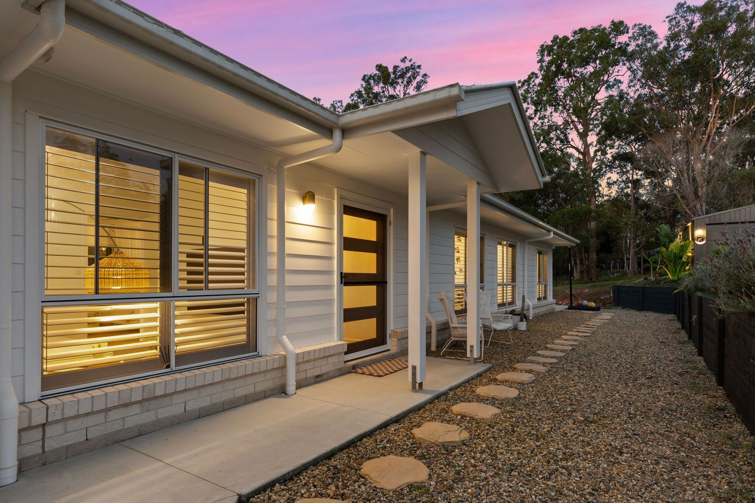 Contemporary home entrance with timber door, stepping stones, louvred windows, and garden at dusk.