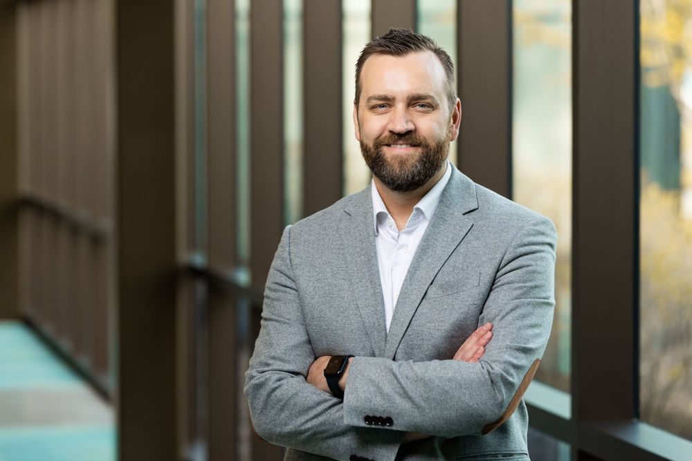 Professional wearing a gray suit and white shirt poses confidently in an office setting.