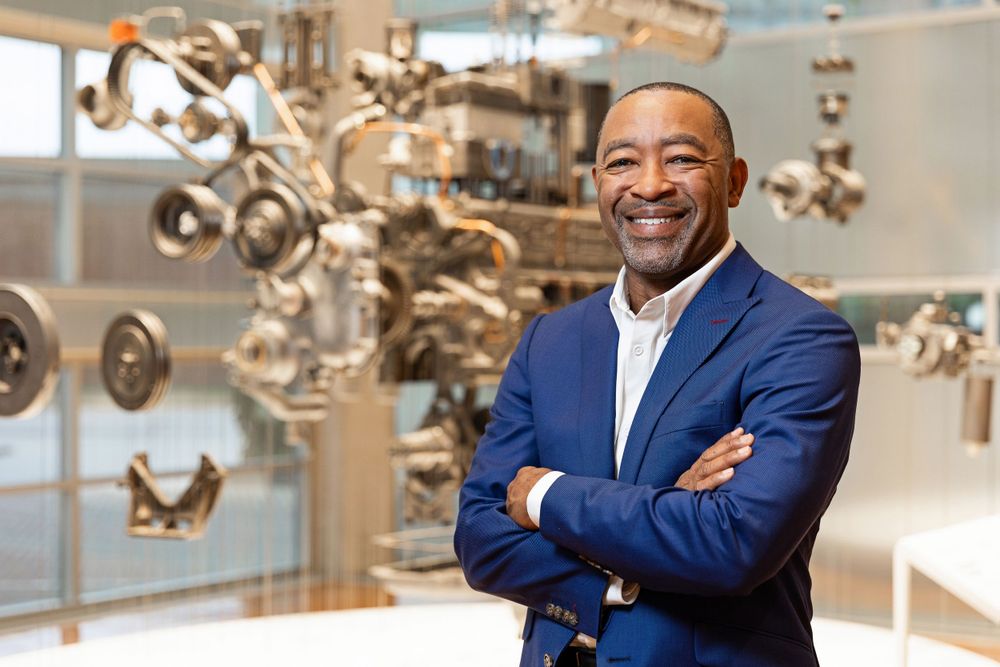 Professional in a navy suit stands confidently in front of technical equipment display.