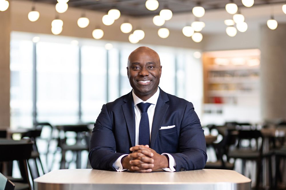 Professional in navy suit sits at table in modern office with pendant lighting.
