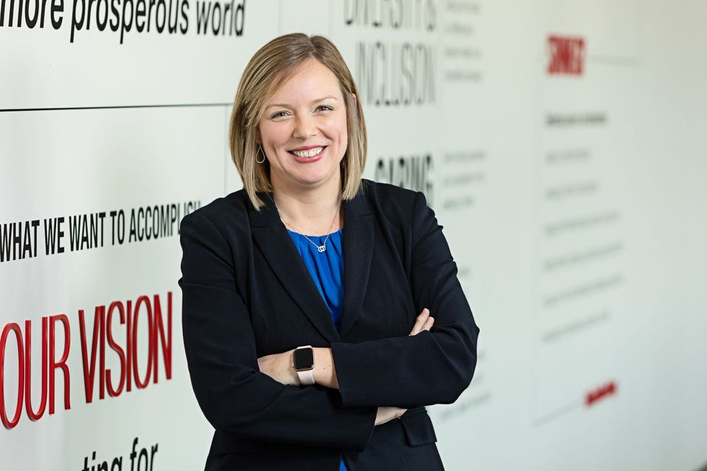 Professional in a dark blazer and blue top poses against a corporate vision statement wall.