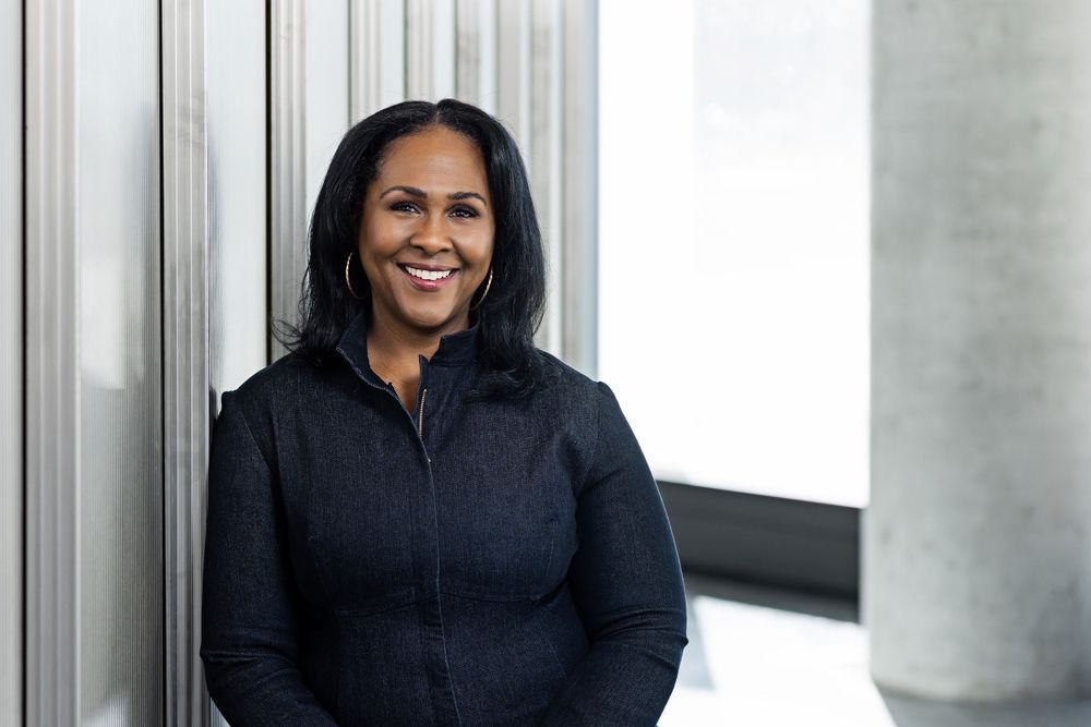 Professional in dark athletic jacket smiles while leaning against modern office column.