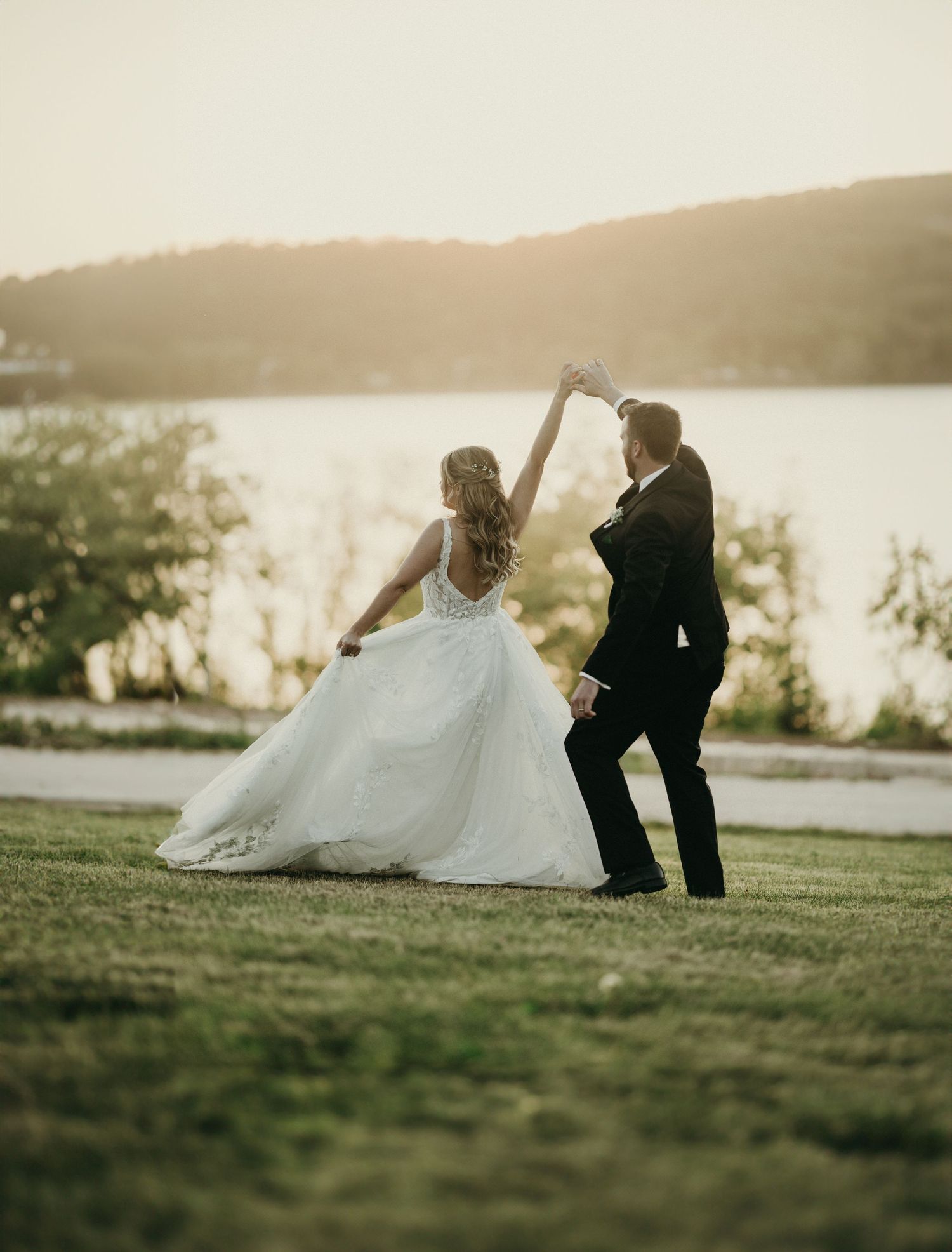 Newlywed couple sits together on grassy hillside overlooking serene lake at sunset in romantic wedding photo series.