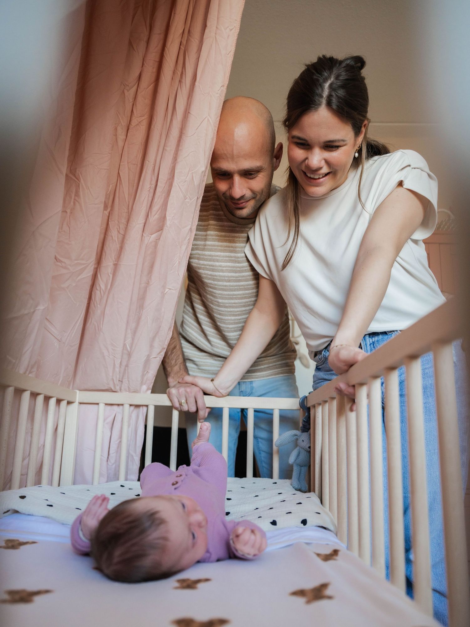 Happy parents smiling over their newborn baby lying in a white crib with pink canopy in nursery.