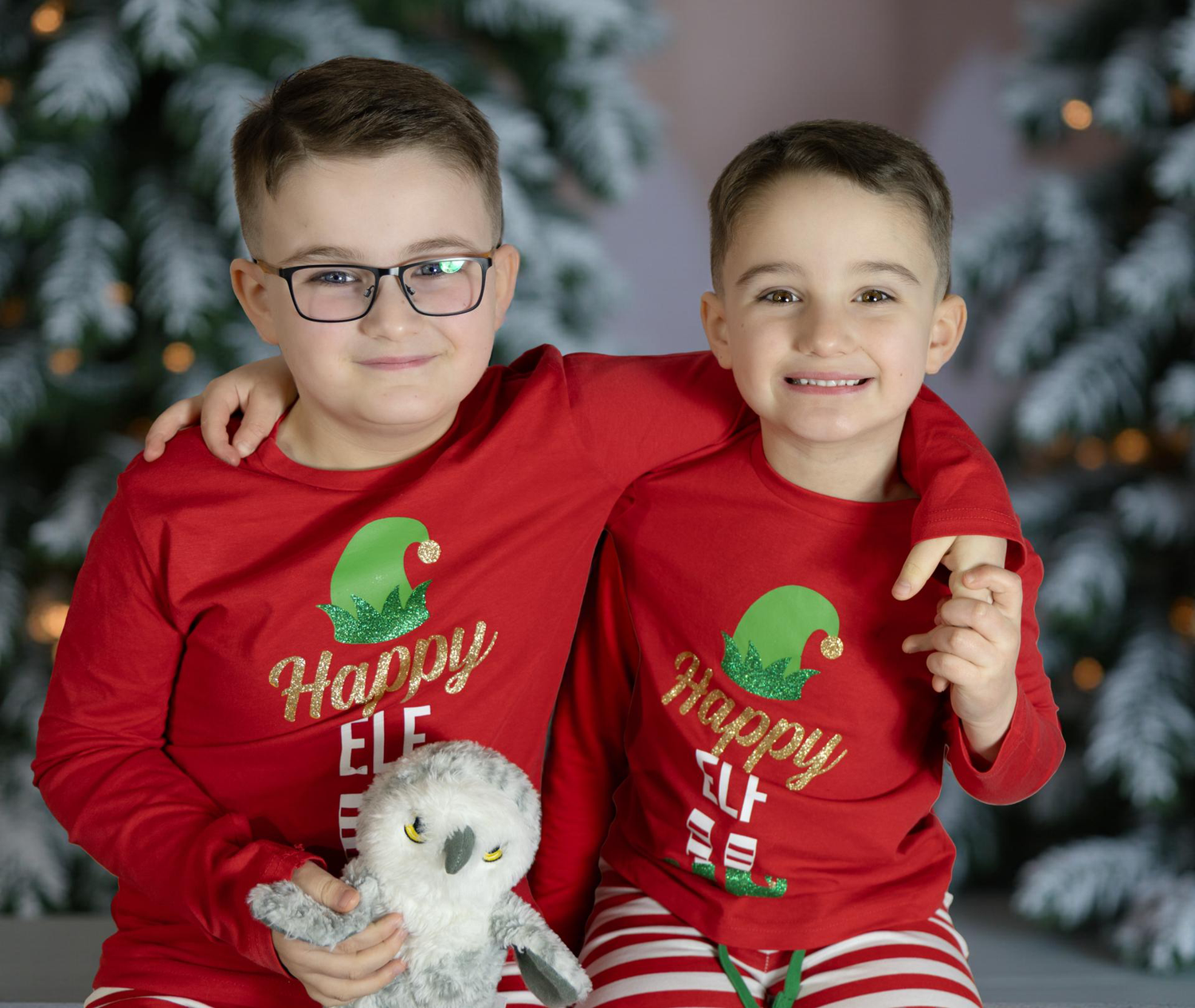 Two children in matching red holiday elf pajamas pose together in front of a Christmas tree.