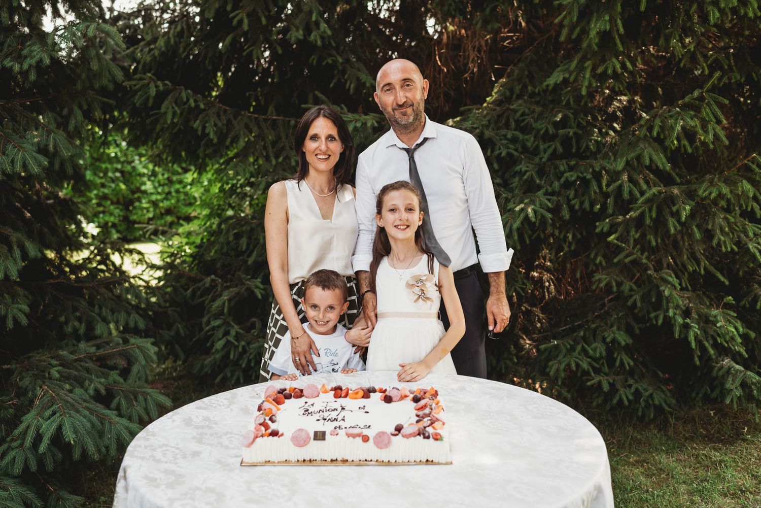 A family stands behind a white decorated celebration cake table in a garden setting.