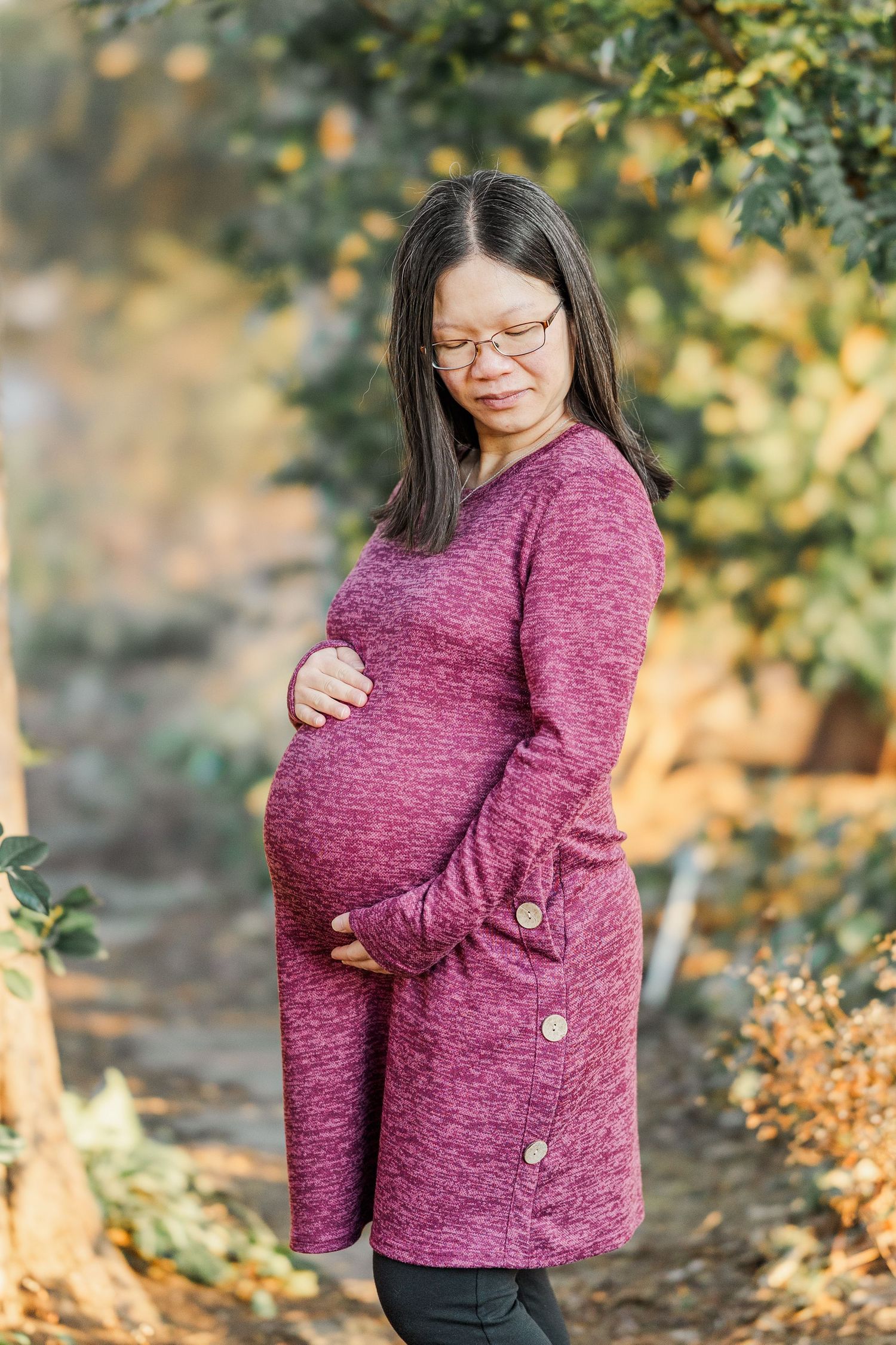 A pregnant person wearing a burgundy sweater poses outdoors in an autumn setting with golden foliage in the background.