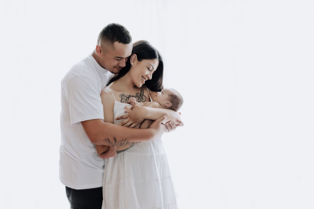 A couple in white outfits shares tender moments with their newborn baby in a bright studio setting.