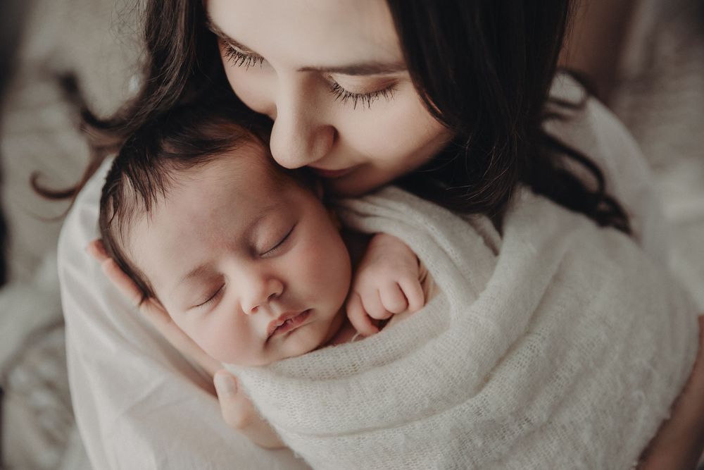 A sleeping baby rests peacefully in a soft white blanket against a neutral background.