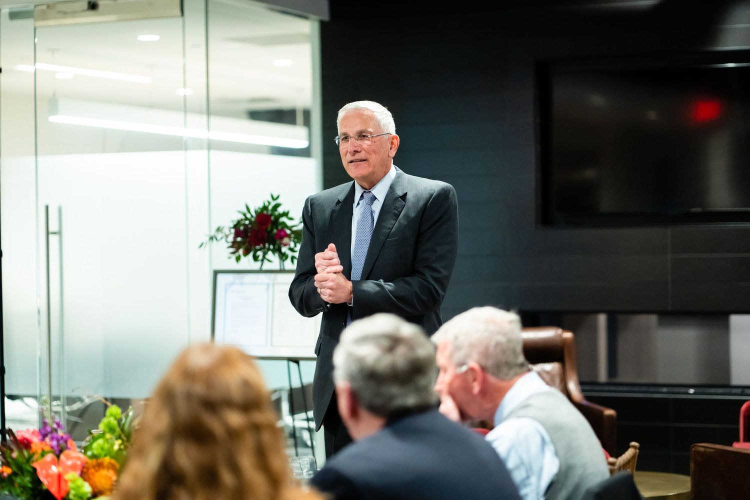 Business people gathered in a modern office conference room with glass walls and colorful floral arrangements.