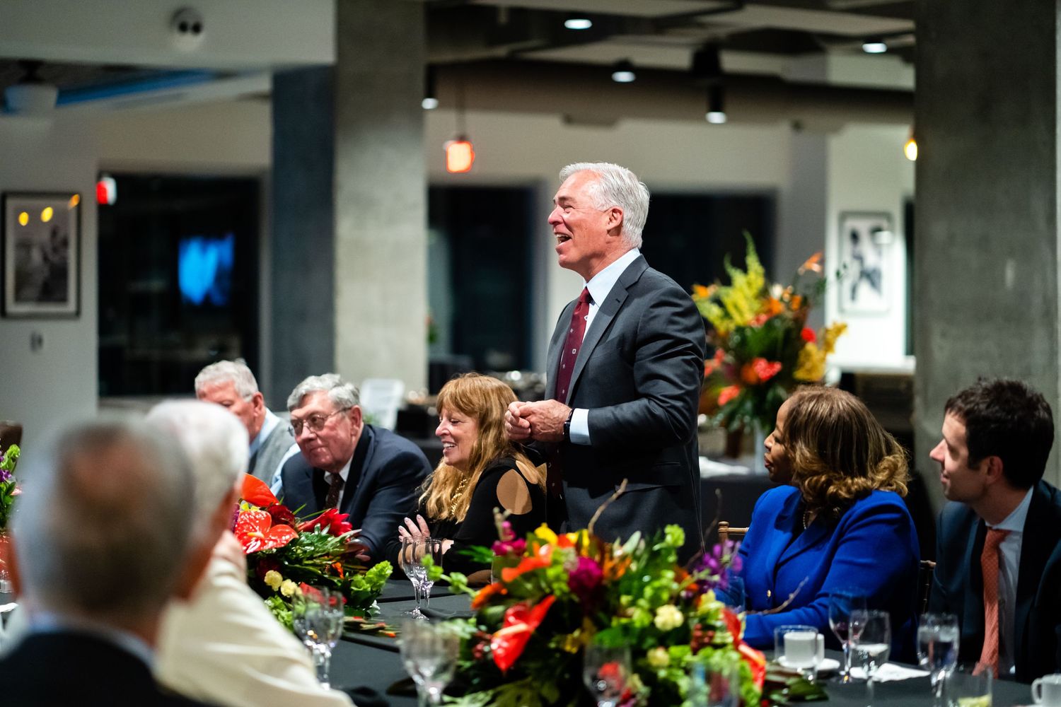 Business professionals seated at a long conference table during a formal dinner event with floral centerpieces.