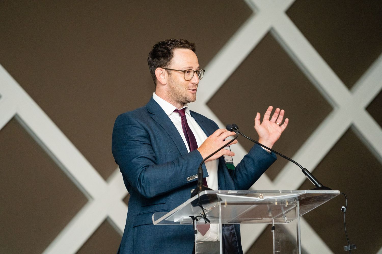 A speaker in a navy suit gestures while giving a presentation at a clear podium on stage.