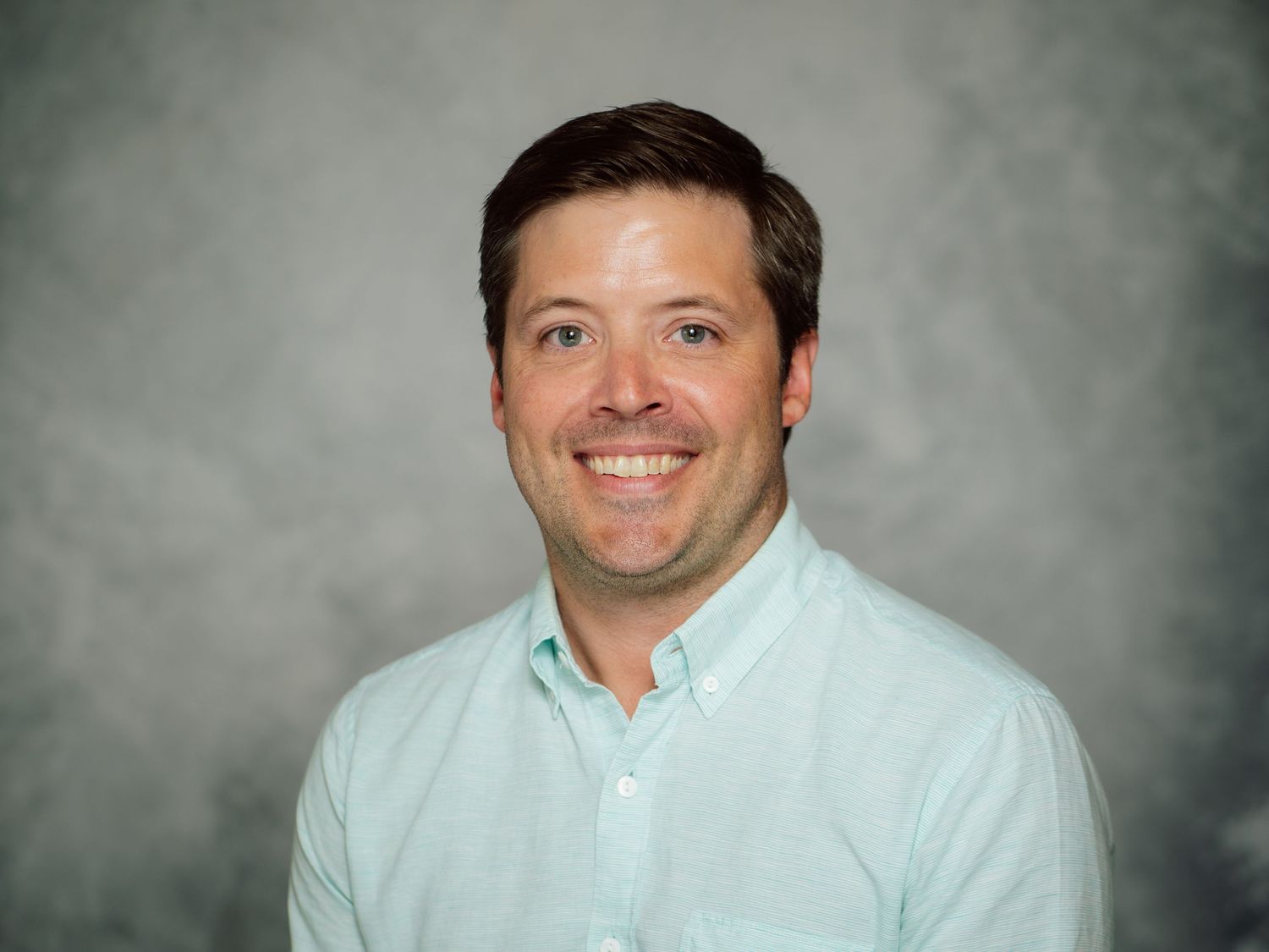 Professional headshot of a person in a light blue button-up shirt against a gray backdrop.