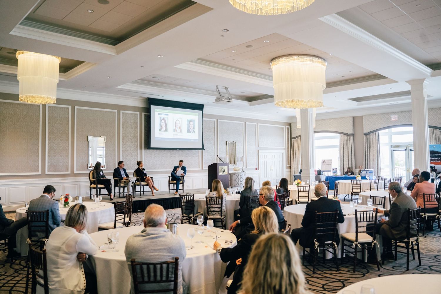 Business conference attendees seated at round tables in an elegant ballroom with crystal chandeliers and presentation screen.