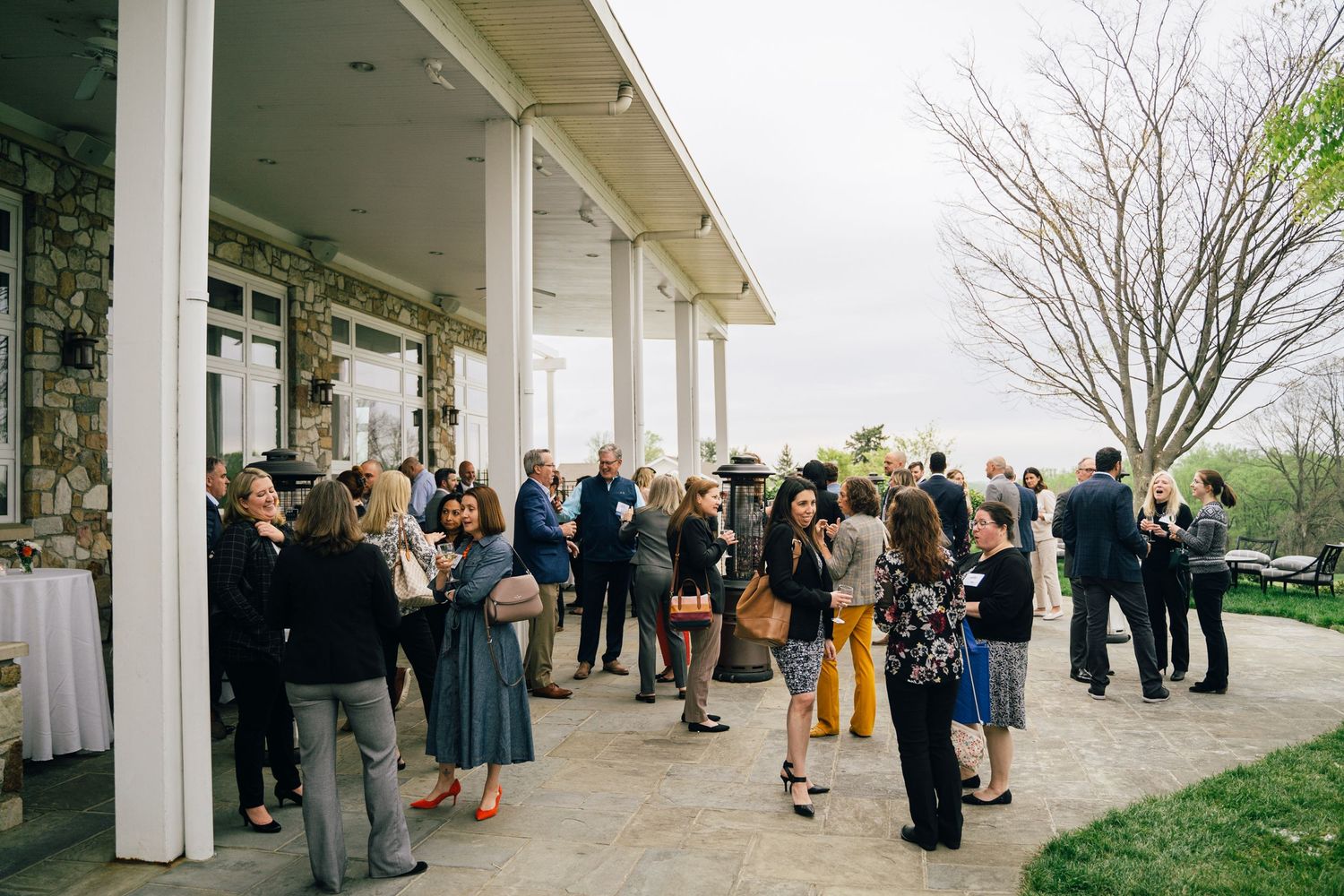 Groups of people gather and network on a white columned porch during an outdoor event.