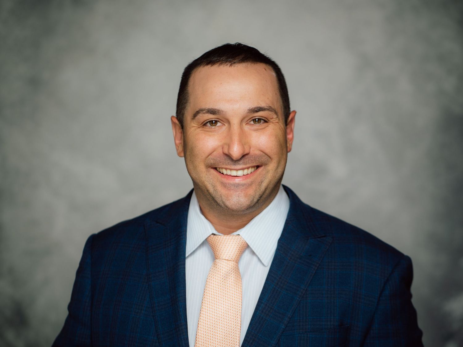 Professional headshot portrait against gray background wearing navy suit and peach tie.