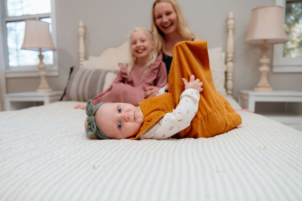 Joyful interaction between mother, daughter and infant on white bedding in bright room. In-home family session.