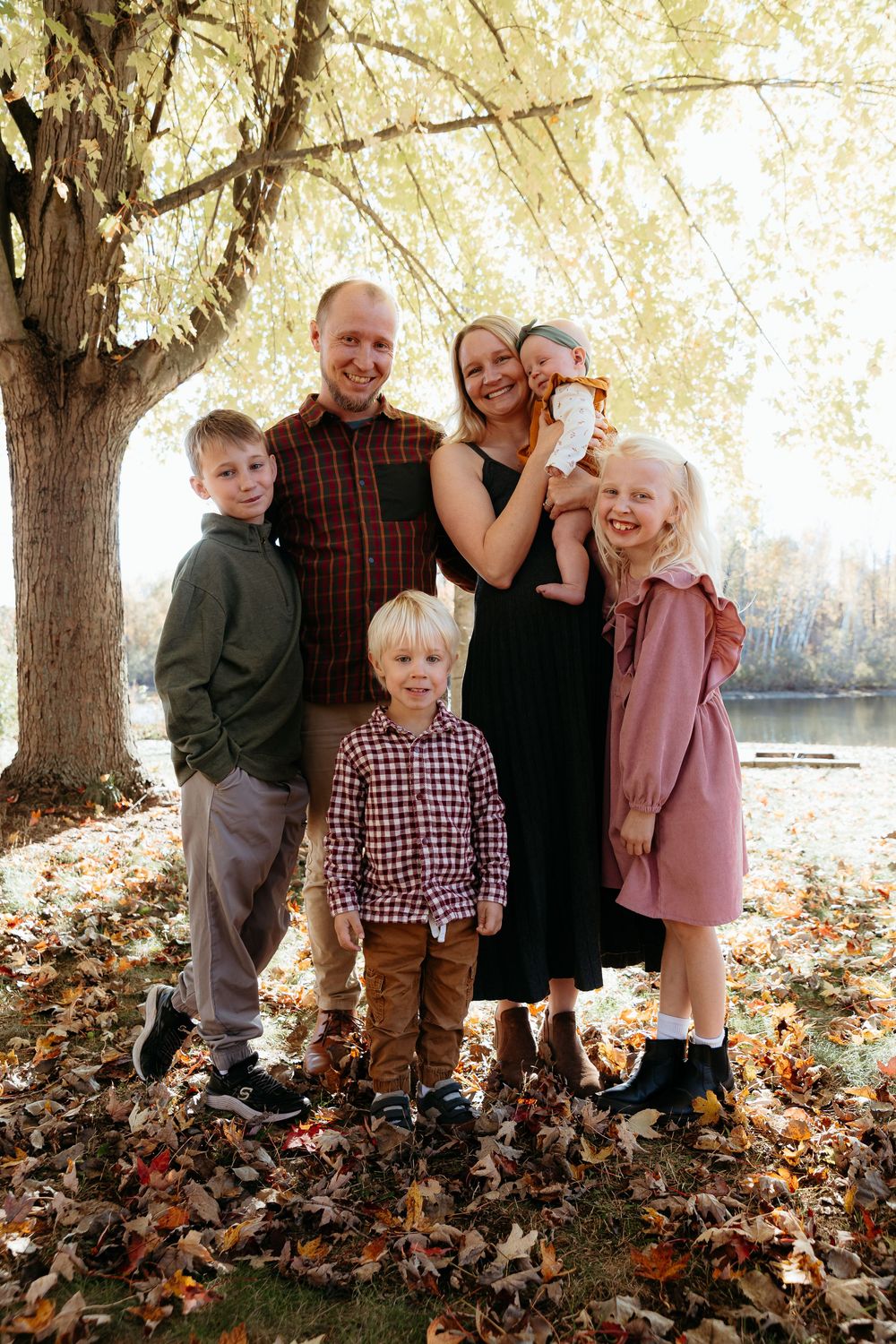 Family group poses together among fallen autumn leaves under large tree branches. Classic family portrait in the fall family photo session.