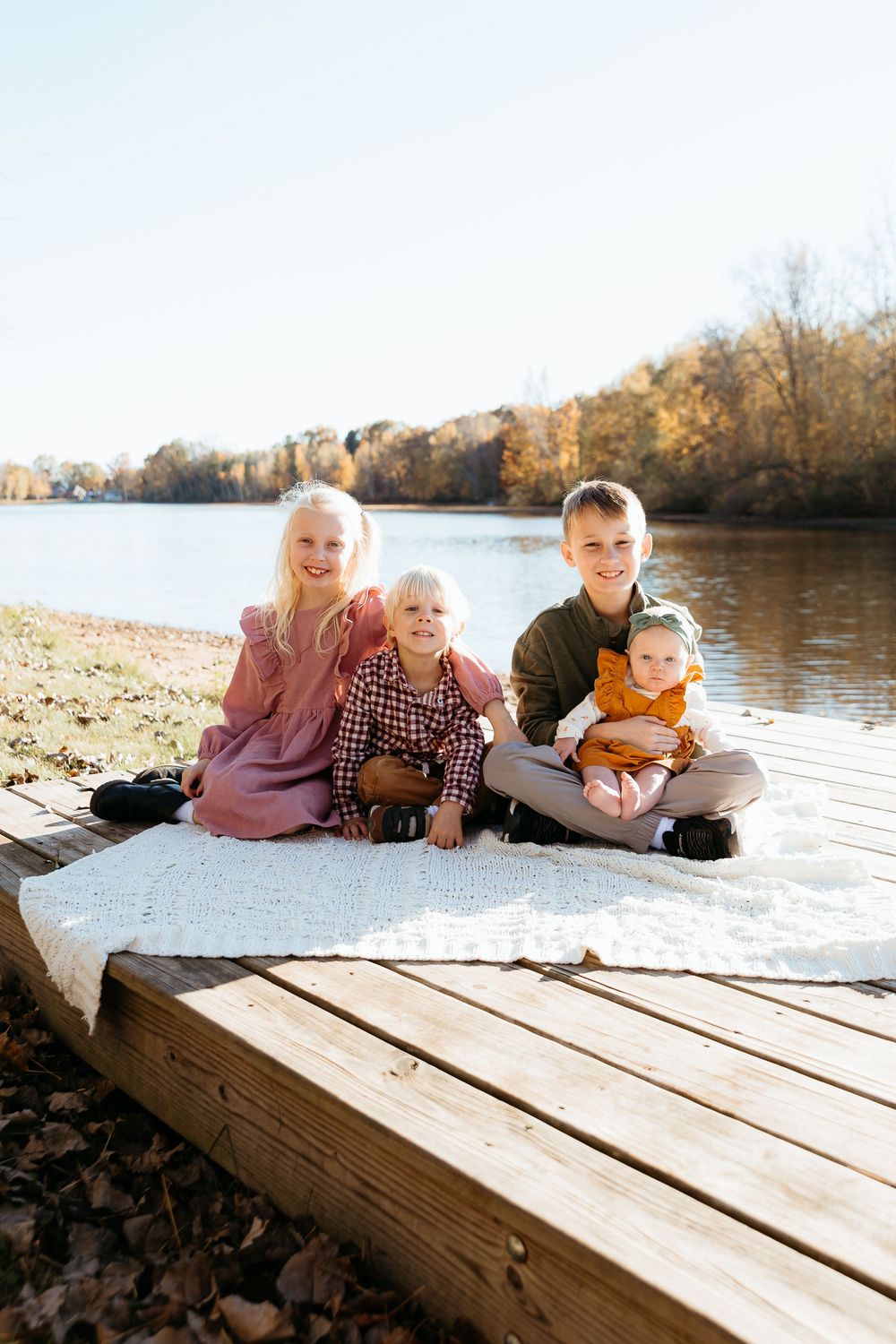 People relaxing on a wooden dock by the water on a sunny fall day. Lakeside family photo session. Children and siblings together.