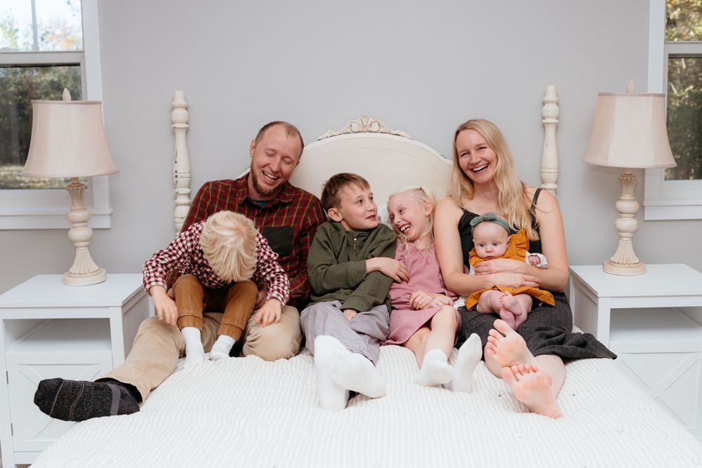 Candid moments of family bonding on a white bed with natural lighting. Laughter and fun at in-home family session. Central Michigan photos