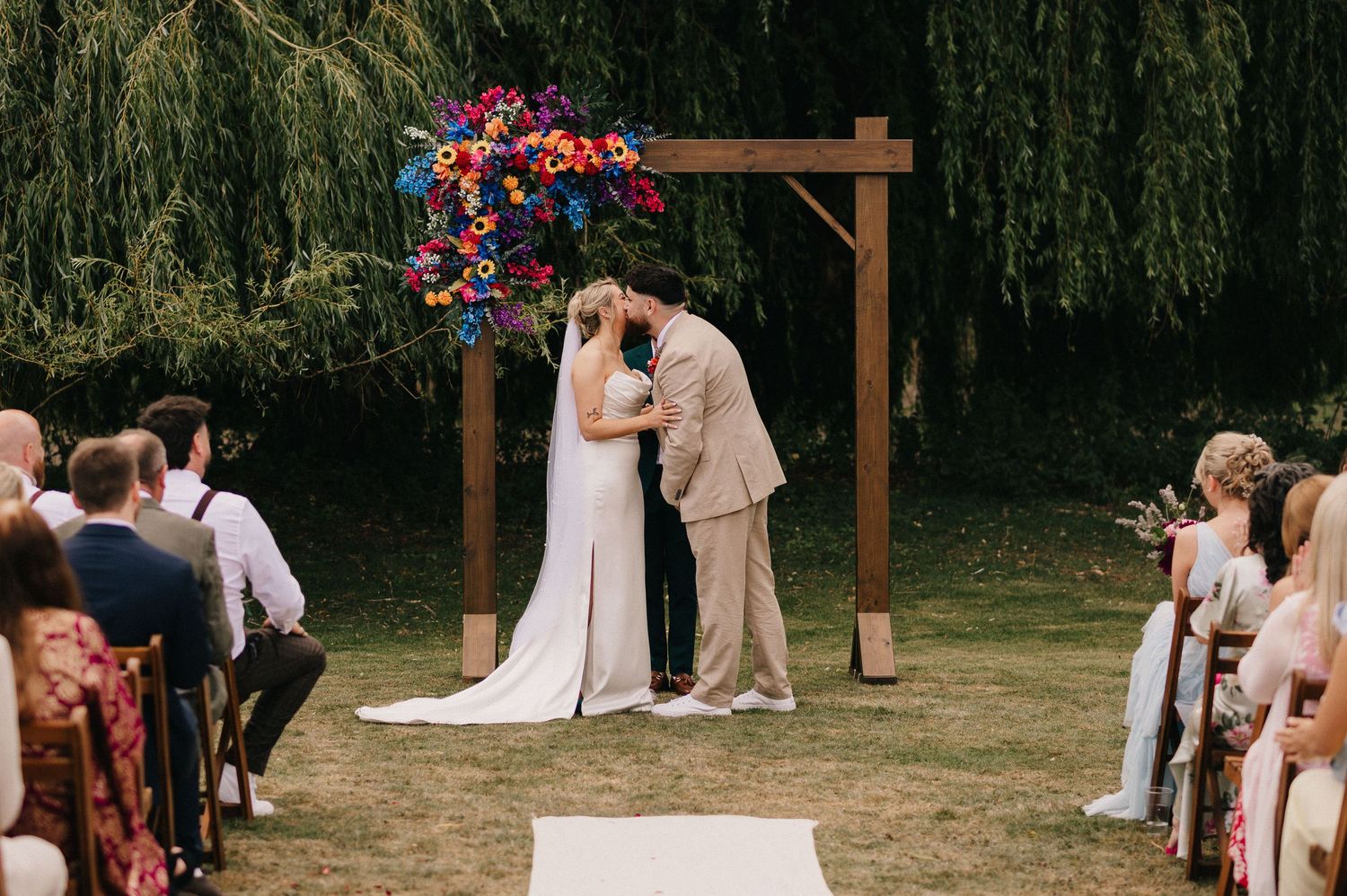 Wedding ceremony under wooden arch with vibrant red and pink floral arrangements in outdoor garden setting.