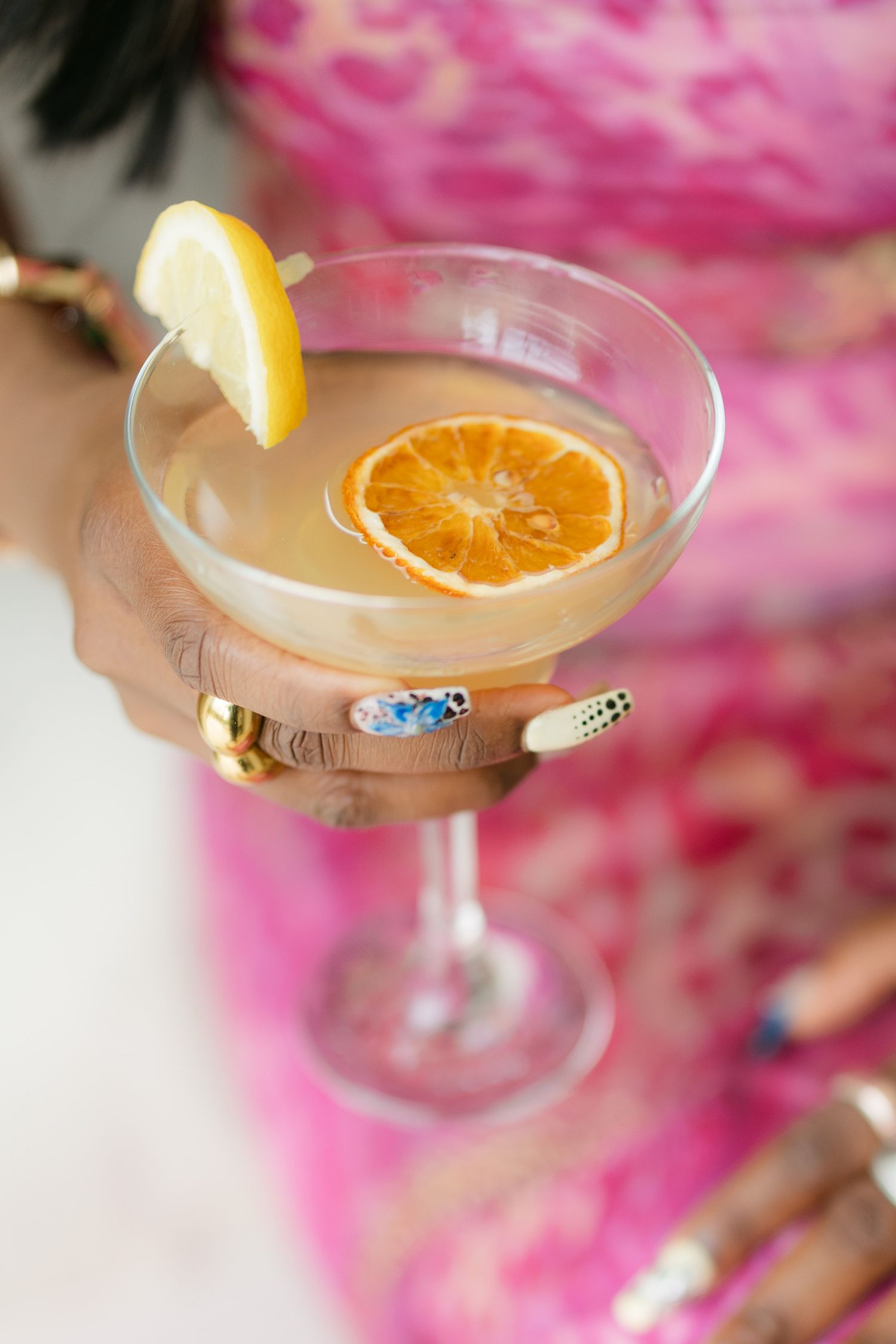 A cocktail glass with citrus garnish held by a hand with decorated nails against a pink background.