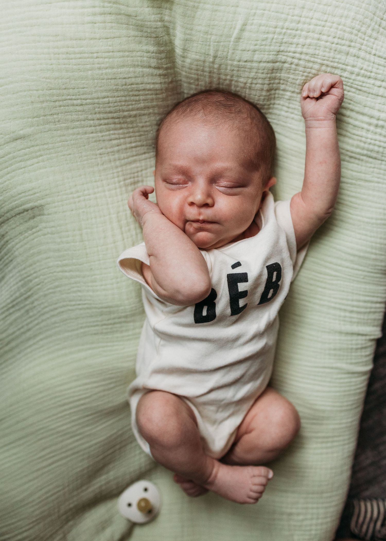 A sleeping baby in a white onesie lies peacefully on a light-colored couch cushion.