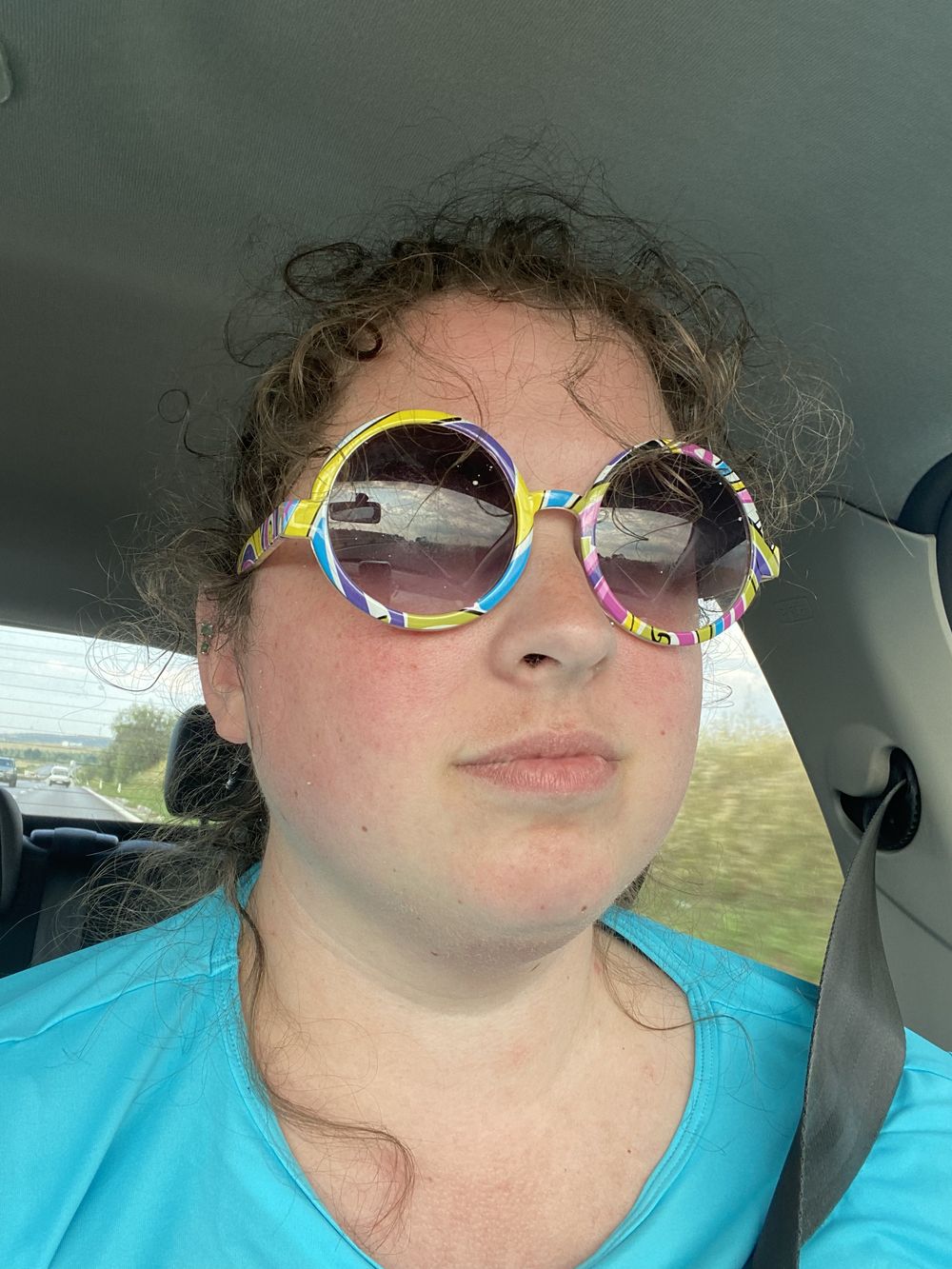 A person wearing colorful sunglasses and a light blue shirt takes selfies while sitting in a car.