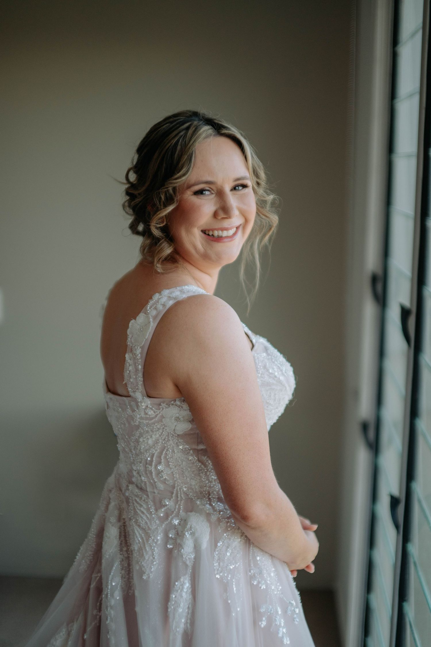 A bride in a white lace wedding dress poses near a window in a series of elegant portraits.