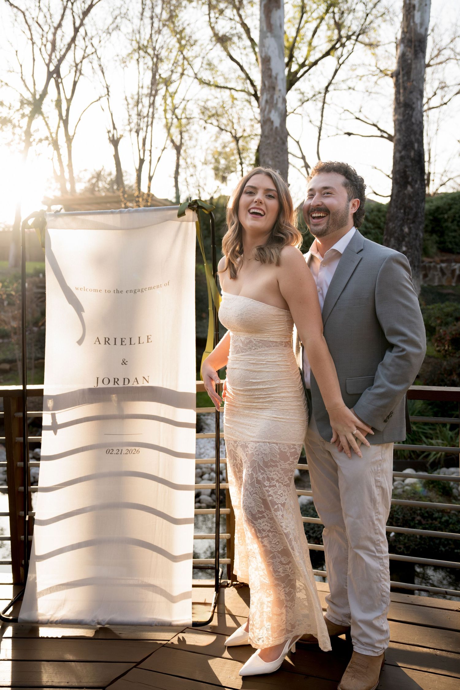 Couple poses with wedding invitation banner in outdoor garden setting.