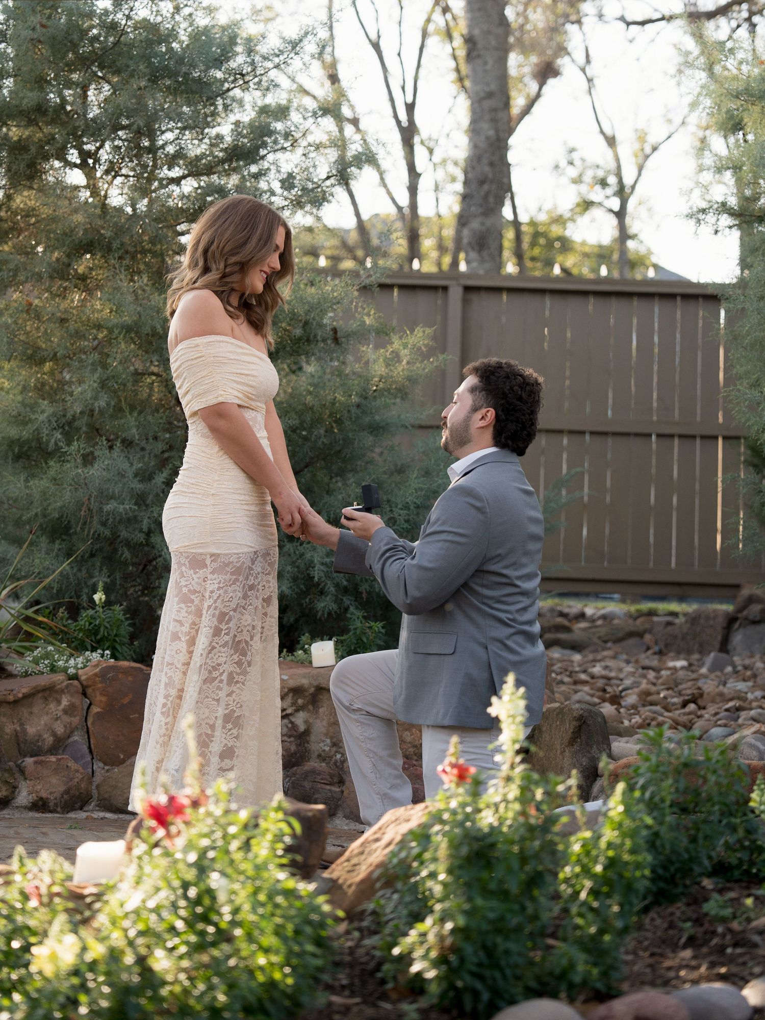 Marriage proposal scene in garden setting with person kneeling among flowering plants.