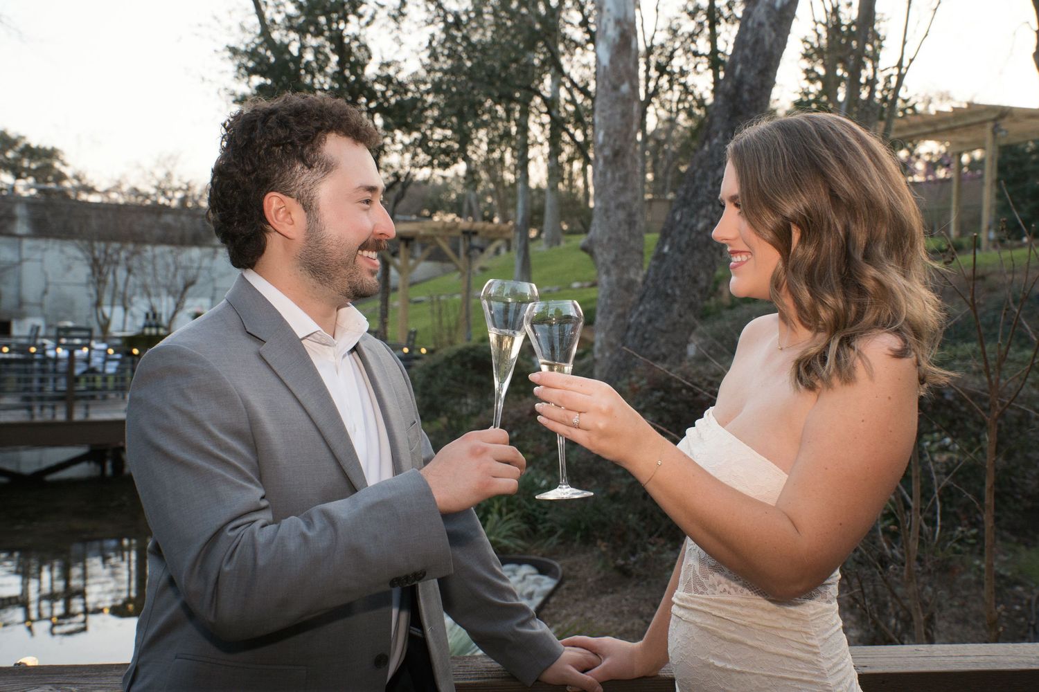 A couple toasts with champagne glasses at an outdoor venue during sunset by a waterfront.