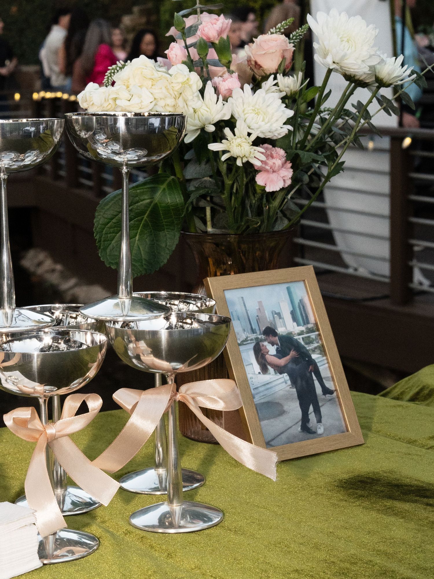 Elegant table setting with champagne glasses tied with pink ribbons, fresh flowers and a framed photo on green tablecloth.
