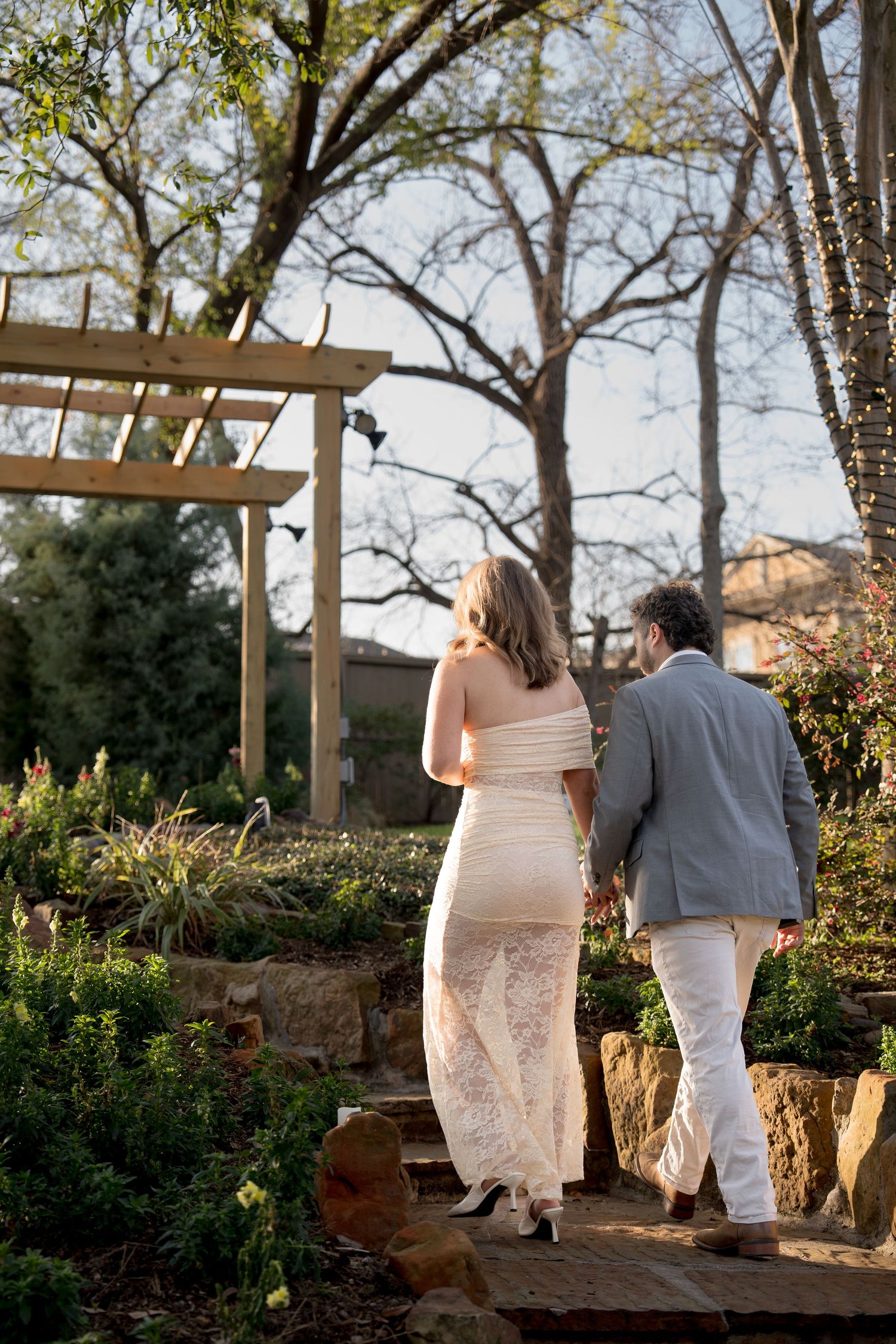 Couple walking together on a garden path at sunset with trees and pergola in background.