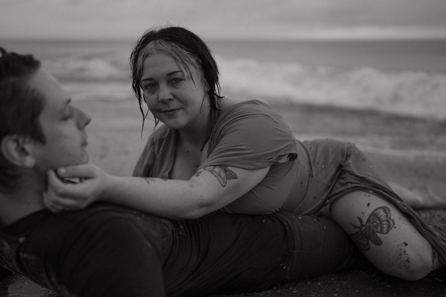 black and white photo of moody, intimate couples session on Cocoa Beach, FL at sunrise. Waves rush over them as golden light comes up.