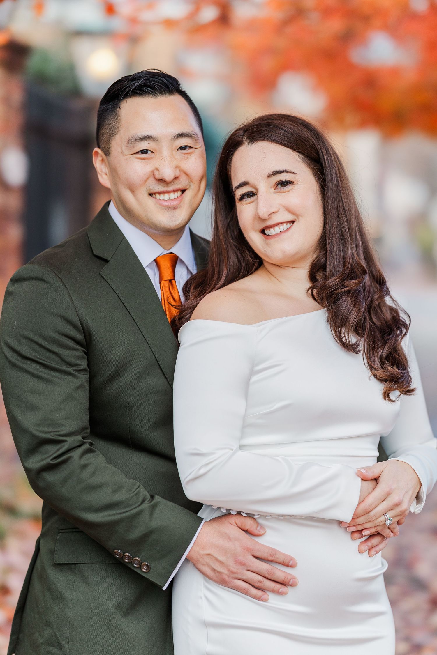 A couple in wedding attire embraces against a backdrop of autumn foliage with orange leaves.
