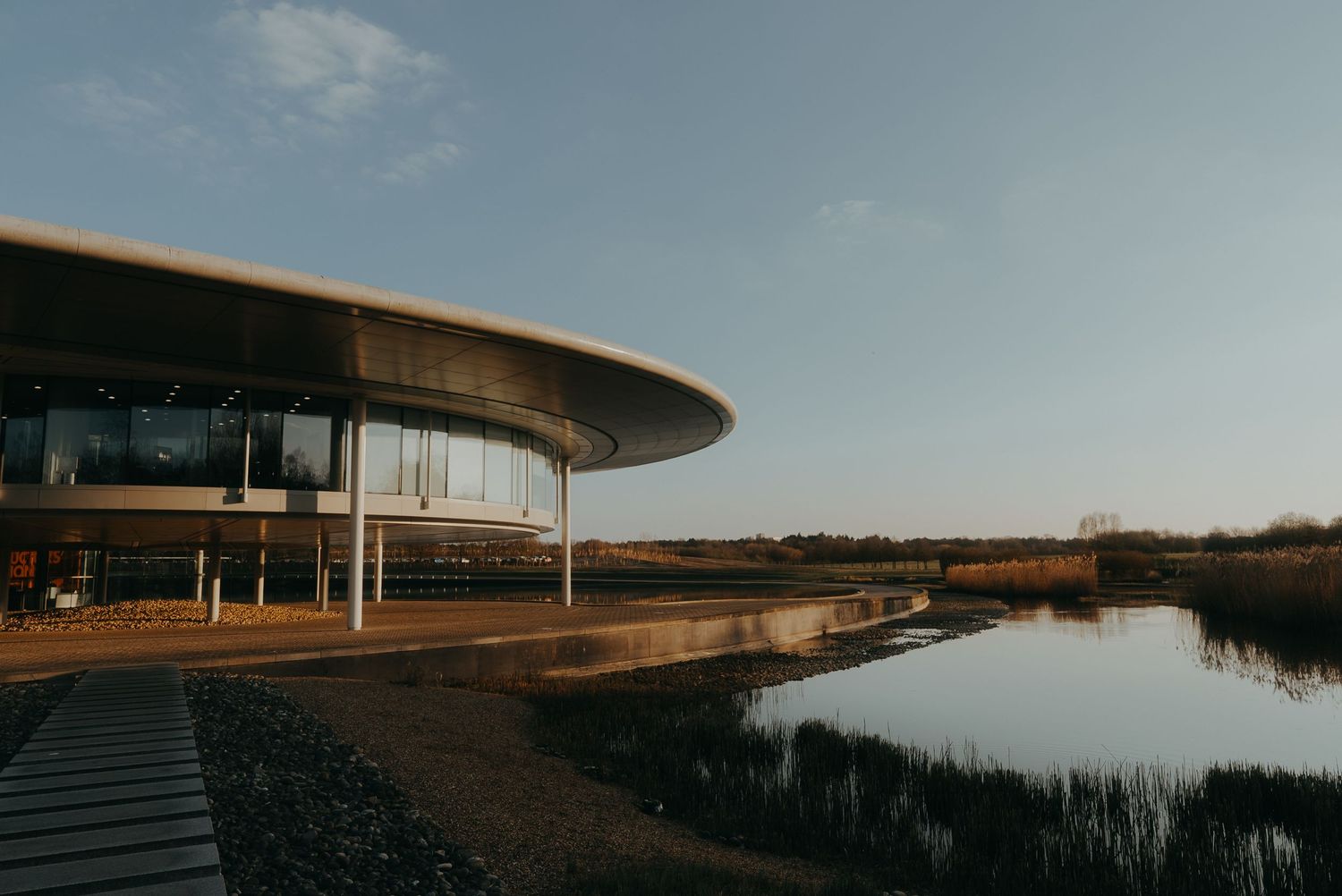 Modern curved building with glass windows reflects sunset light against a serene waterfront landscape.