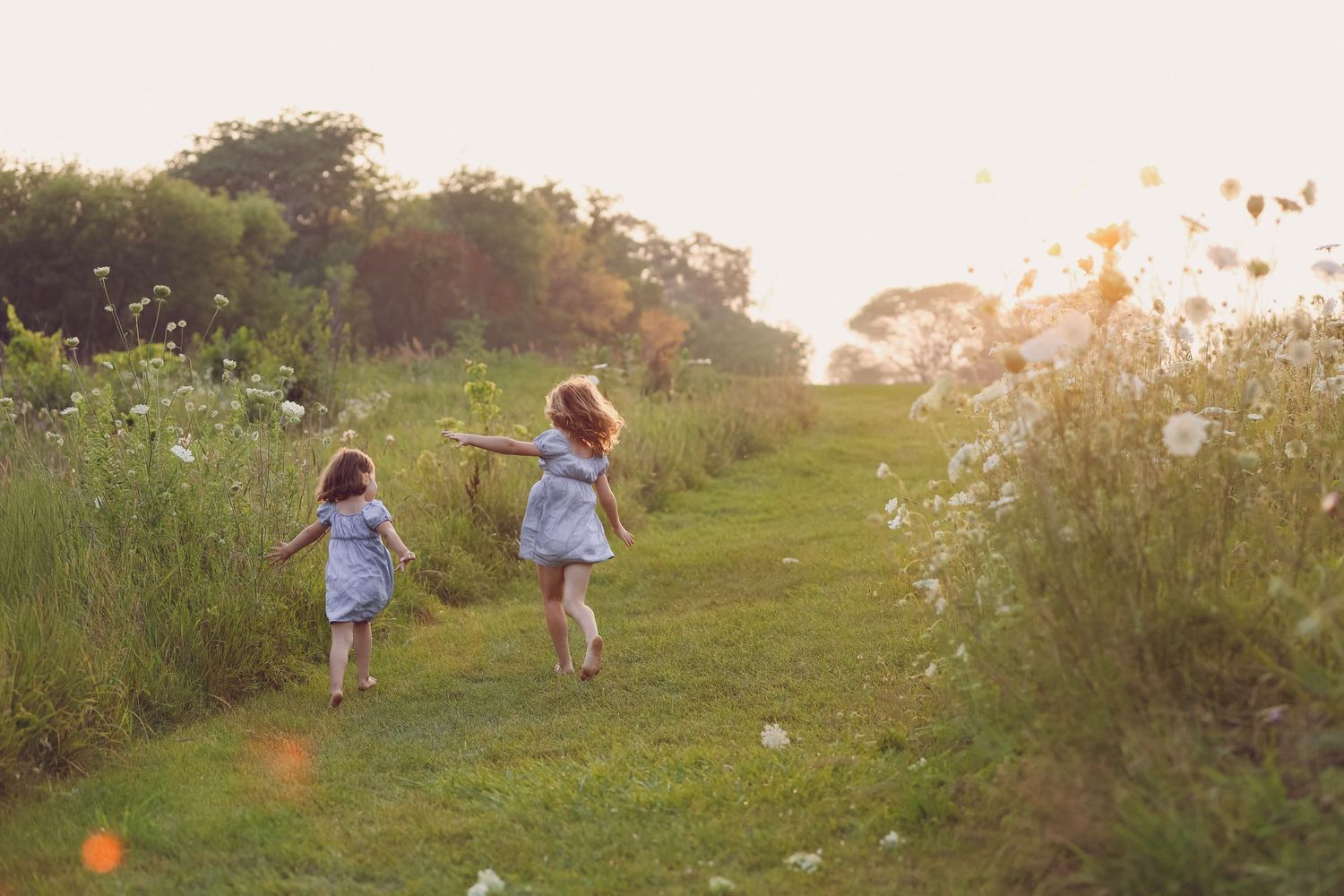 Two young girls in blue dresses run through a sunlit wildflower meadow path at golden hour.