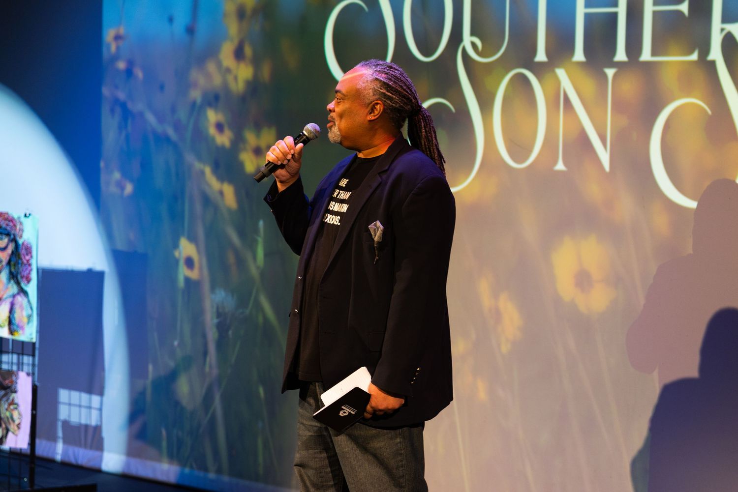 A performer in a black jacket sings on stage with a microphone against a blue-lit backdrop with text display.