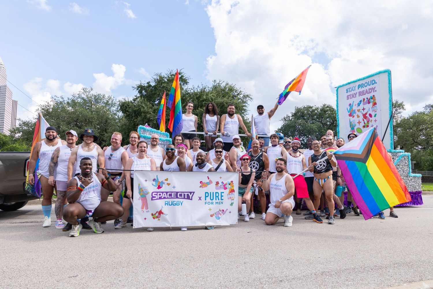 A group of people wearing white shirts gather outdoors holding rainbow pride flags and banners at a community event.