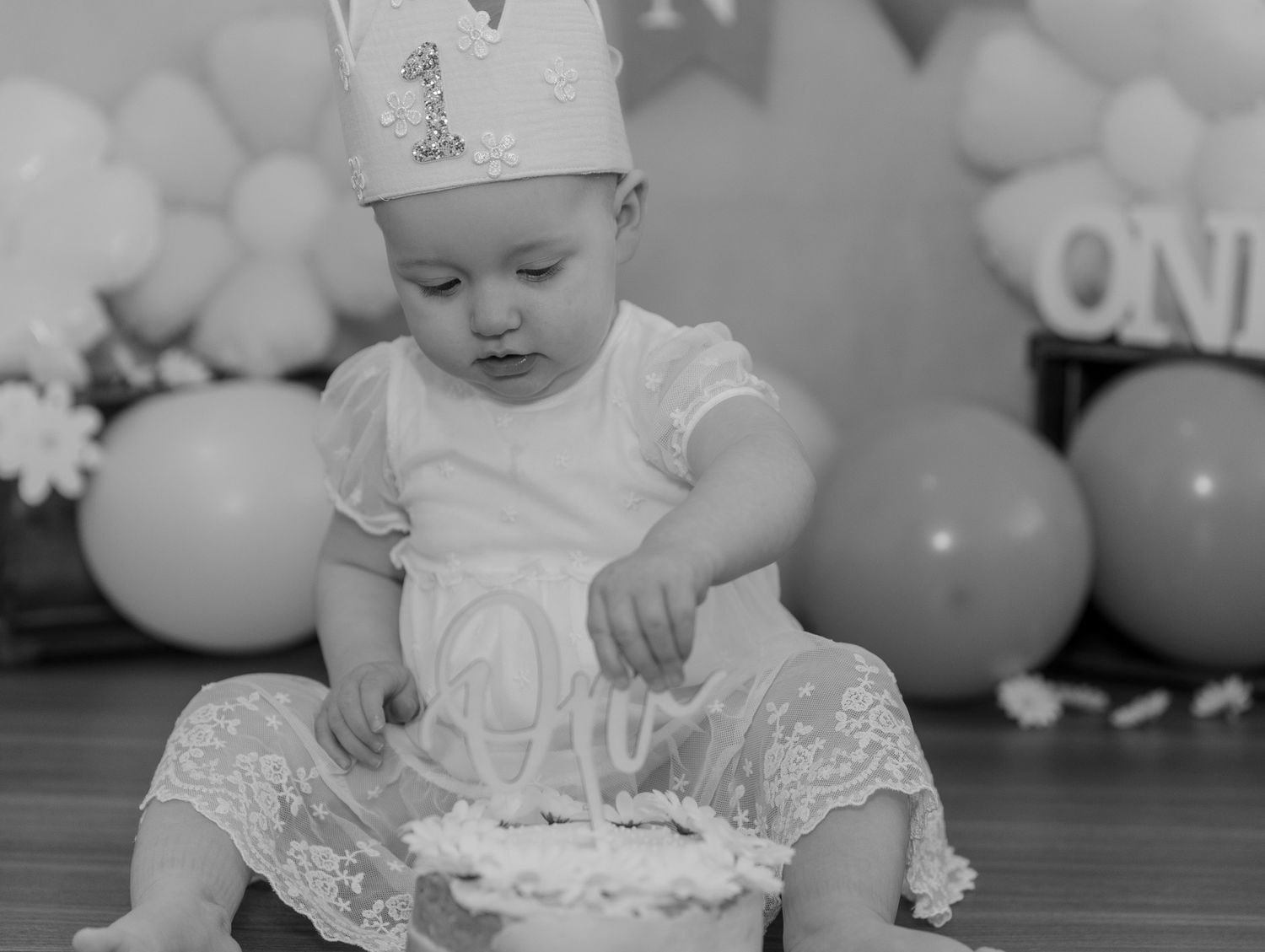 A baby wearing a white outfit explores their first birthday cake during a cake smash photo session.