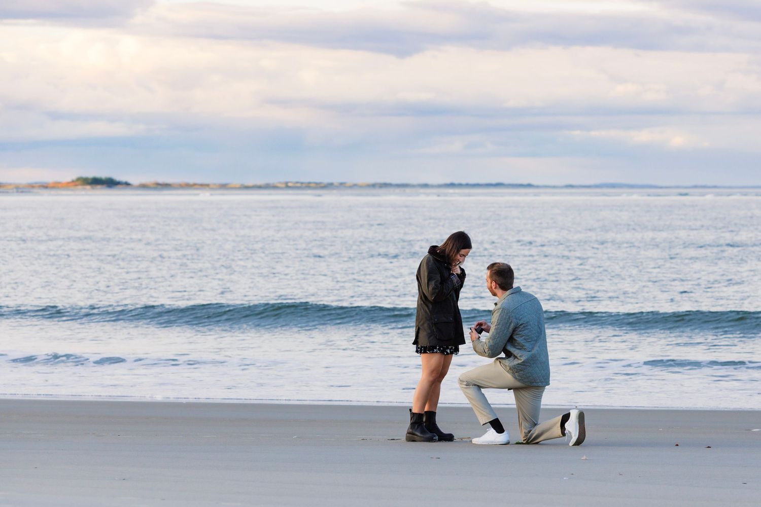 Couple kneeling on a sandy beach during a romantic proposal at sunset by the ocean waves.