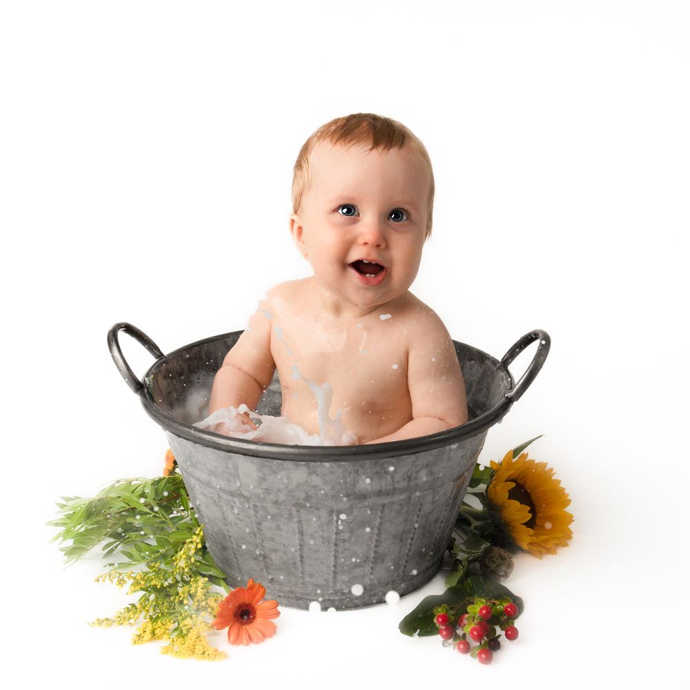 A happy baby sits in a rustic galvanized tub with bubbles, surrounded by colorful flowers on a white background.